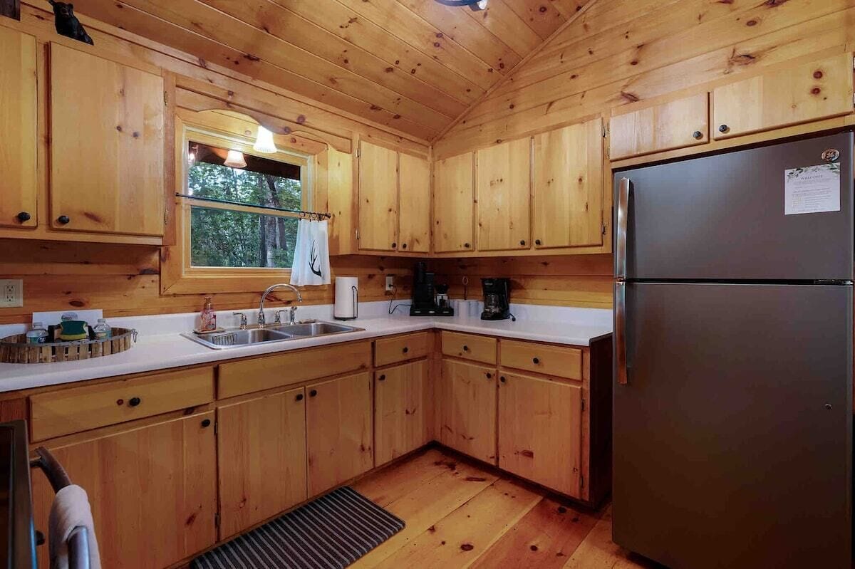 Une cuisine rustique aménagée dans une cabane en bois, avec un réfrigérateur en acier inoxydable, des placards en bois, une fenêtre offrant une belle vue et un évier équipé d’une bouilloire.
