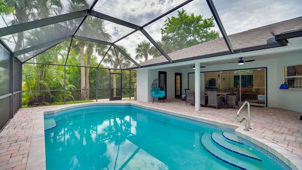 A swimming pool with a glass enclosure, surrounded by a patio area with outdoor furniture and a view of palm trees.