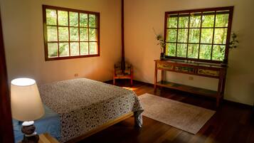 Une chambre avec un lit, un bureau, une lampe et deux fenêtres donnant sur un paysage verdoyant.