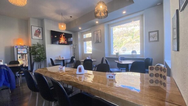 Um restaurante com uma longa mesa de madeira, cadeiras pretas, luminárias pendentes e uma televisão exibindo uma lareira.