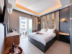 A modern hotel bedroom with a large bed, a wooden side table with a bottle and glasses, a TV, and a balcony with a view of the city.