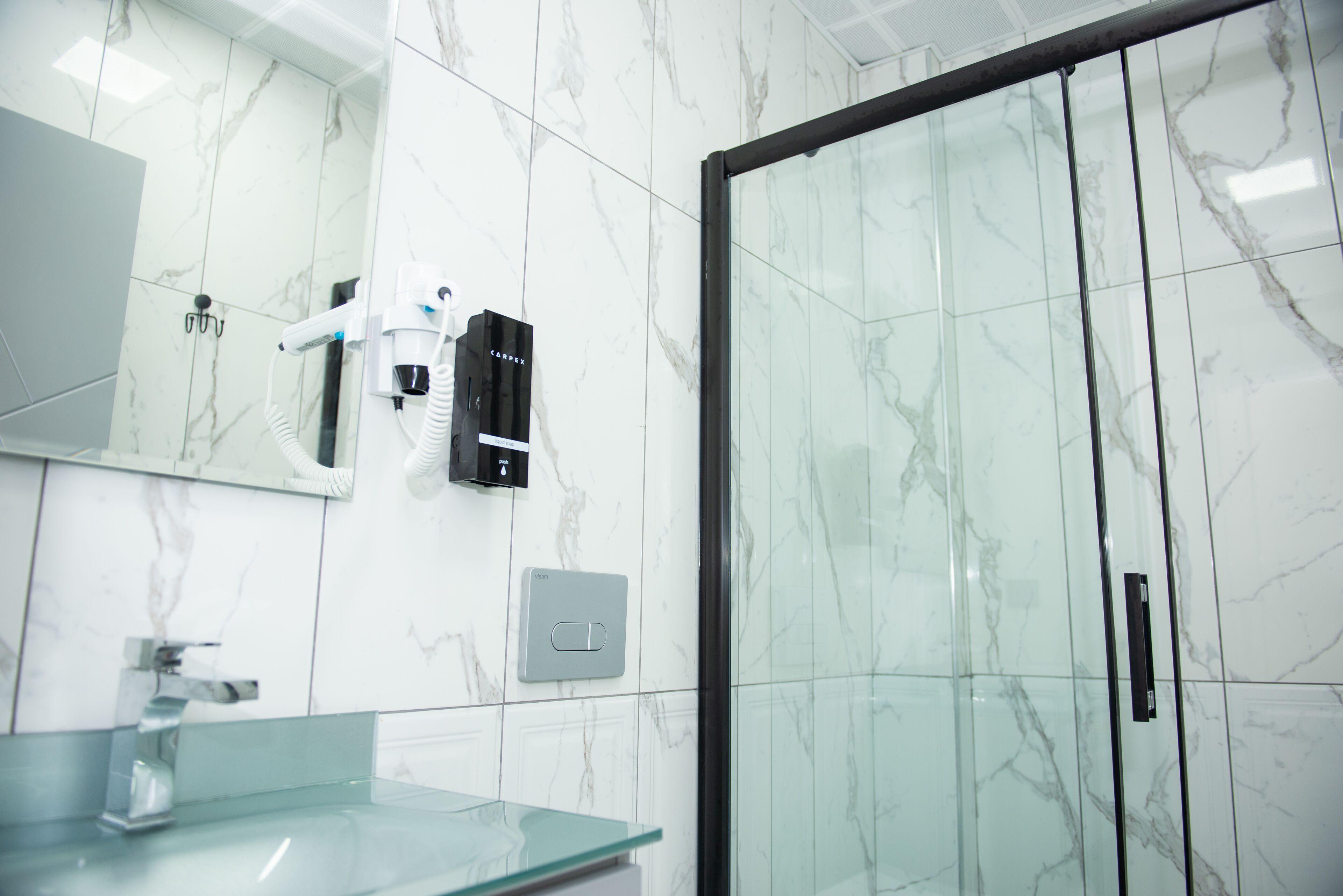 Un baño con cabina de ducha de vidrio, pared con azulejos de mármol, lavamanos de vidrio y un secador de pelo montado en la pared.