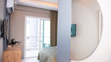 A modern hotel room with a wooden desk, a bed with a patterned bedspread, a television mounted on the wall, and a balcony with a view.