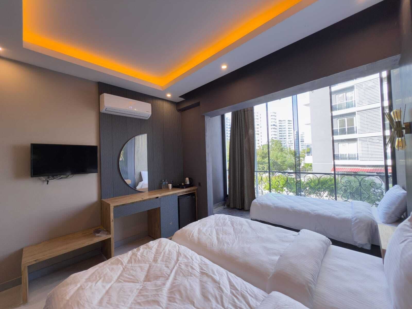 A modern hotel room with two beds, a TV, a mirror, and a large window with a view of buildings.