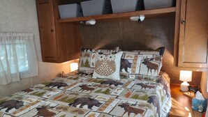 A bedroom with a bed featuring a wildlife-themed quilt, wooden headboard, built-in storage, bedside lamps, and a window with sheer curtains.
