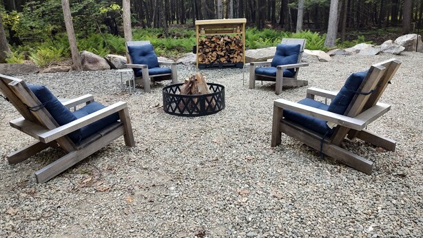 A circular seating arrangement with four lounge chairs around a central fire pit, set on a gravel surface with a forest backdrop.