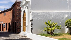 Una calle con un edificio blanco que tiene una puerta de madera y un pequeño jardín con plantas de plátano.