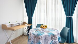 Una habitación compacta con un escritorio de madera, una mesa con frutero, cortinas color turquesa y vistas a través de cortinas blancas traslúcidas.
