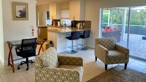 A modern living room with a glass dining table, two patterned armchairs, a black office chair, and a kitchen area with bar stools.