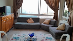 A living room with a sofa, a TV on a wooden cabinet, and a view of the outdoors through large windows. - Authentic Lebanon - Charming, welcoming property in the heart of Magdouché (Maghdoucheh)