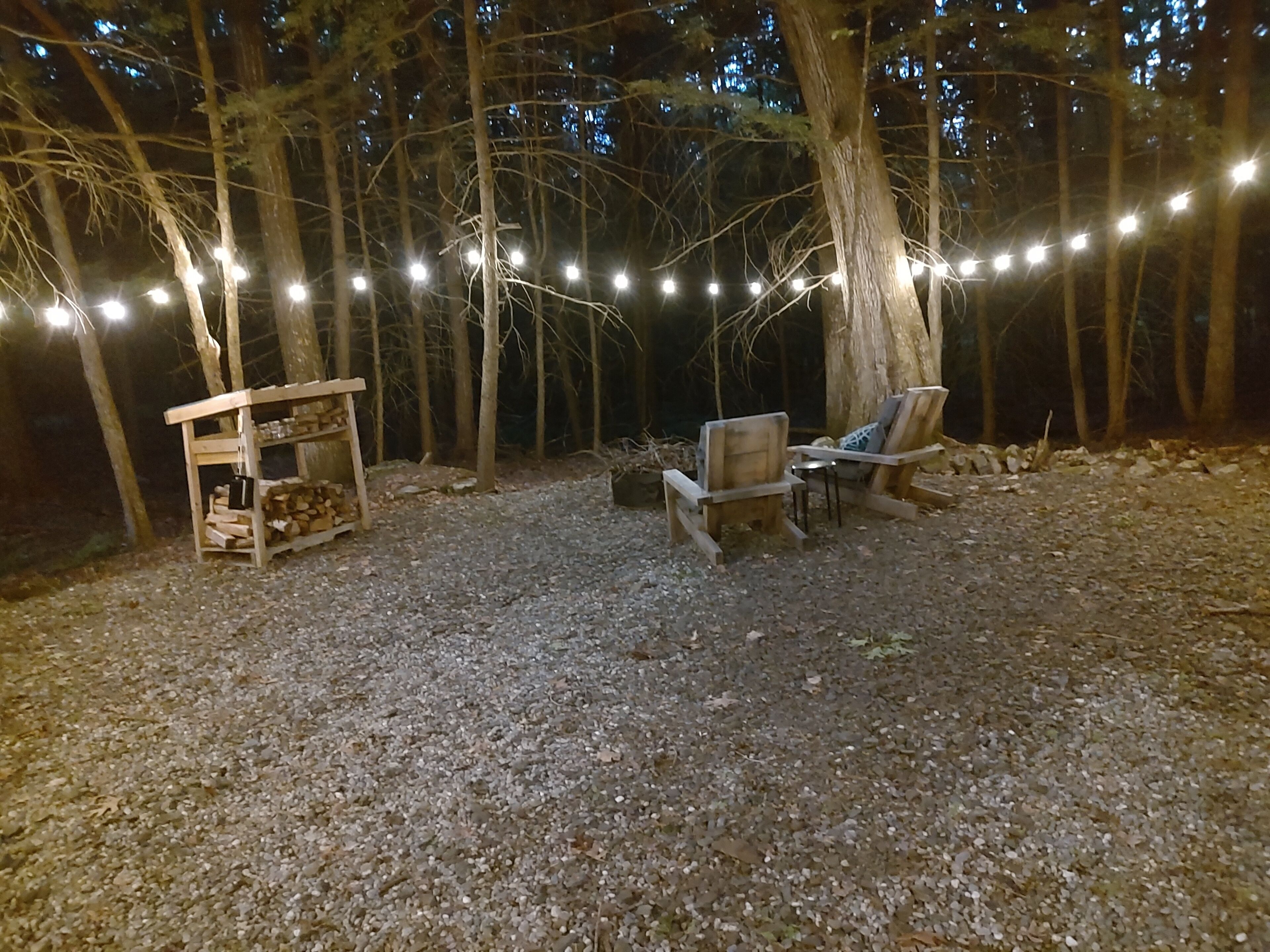 Ein rustikaler Sitzbereich im Freien mit zwei hölzernen Stühlen und einem kleinen Holztisch, umgeben von Bäumen und Lichterketten.