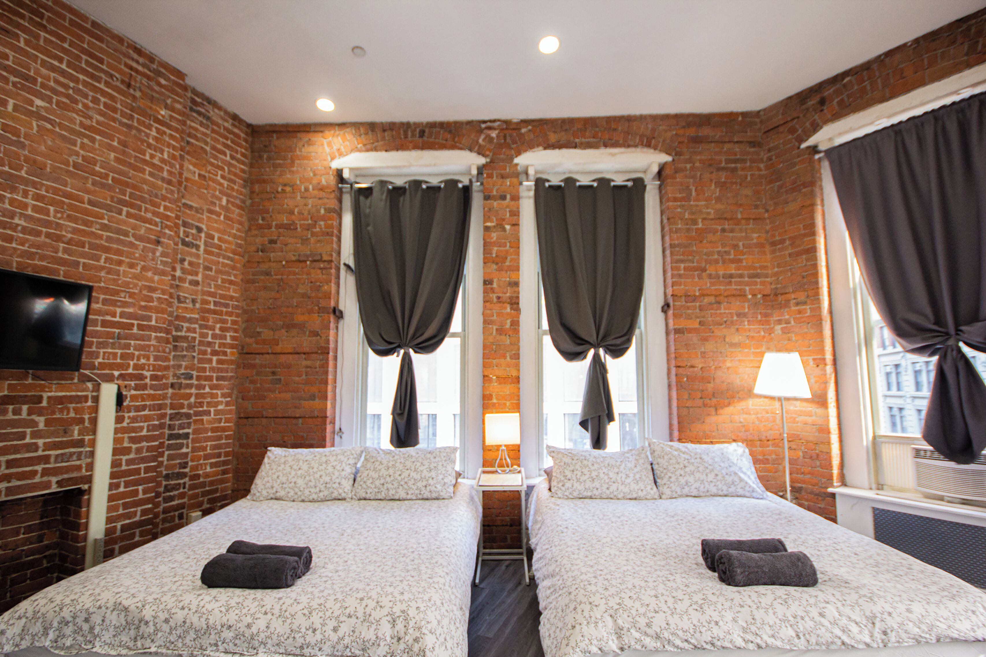 A hotel room with two beds, a flat-screen TV, a brick wall, and a window with curtains.
