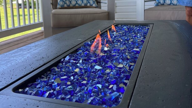Ein Tisch mit Glasplatte, blauem Glas und darunter orangefarbenen Flammen, auf einer Veranda mit zwei beigefarbenen Stühlen und einem dekorativen Wandzeichen.
