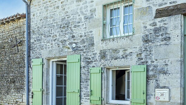 Ein Steinhaus mit zwei Fenstern, jedes mit grünen Fensterläden, und einer kleinen Tür.