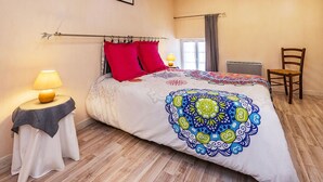 Ein Schlafzimmer mit Bett, Sessel, Nachttisch mit Lampe, Fenster mit Vorhängen und einem gerahmten Bild an der Wand.