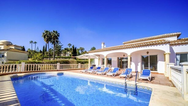 Une piscine entourée de chaises longues, un bâtiment blanc orné d’arches et, au loin, des palmiers.