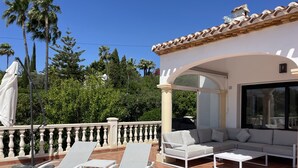 Un espace extérieur aménagé, partiellement abrité, avec des fauteuils de jardin blancs et un canapé gris, entouré d’une rambarde blanche et offrant une vue sur des palmiers et une végétation luxuriante.