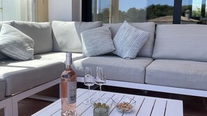 Un espace extérieur ensoleillé, aménagé avec une table blanche, des coussins et une bouteille de vin.