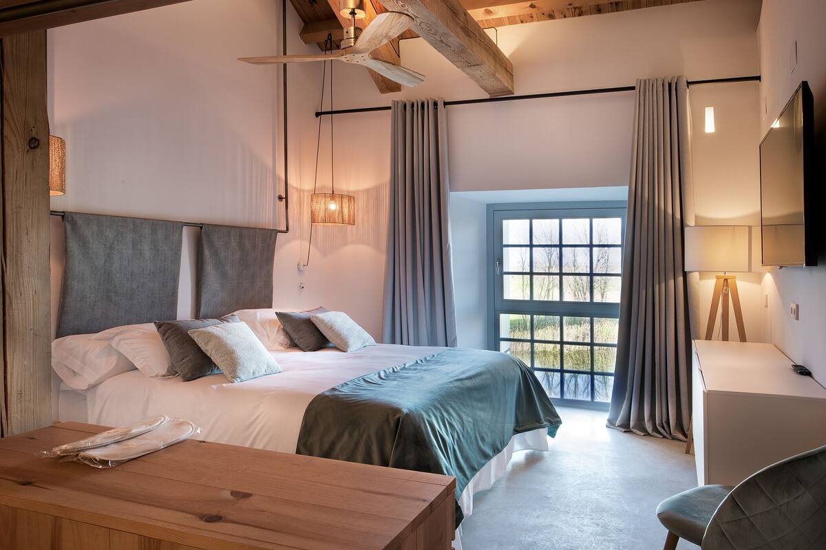 A modern bedroom with a wooden ceiling, a large bed with a grey and white duvet, a wooden bedside table, a desk, a chair, and a window with curtains.