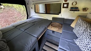 Interior compacto de una caravana con dos zonas de descanso, una mesita pequeña y un estante con libros y una planta.