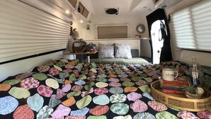 Un dormitorio pequeño y bien iluminado, con un edredón colorido, una mesita de noche con libros y una ventana con persianas.