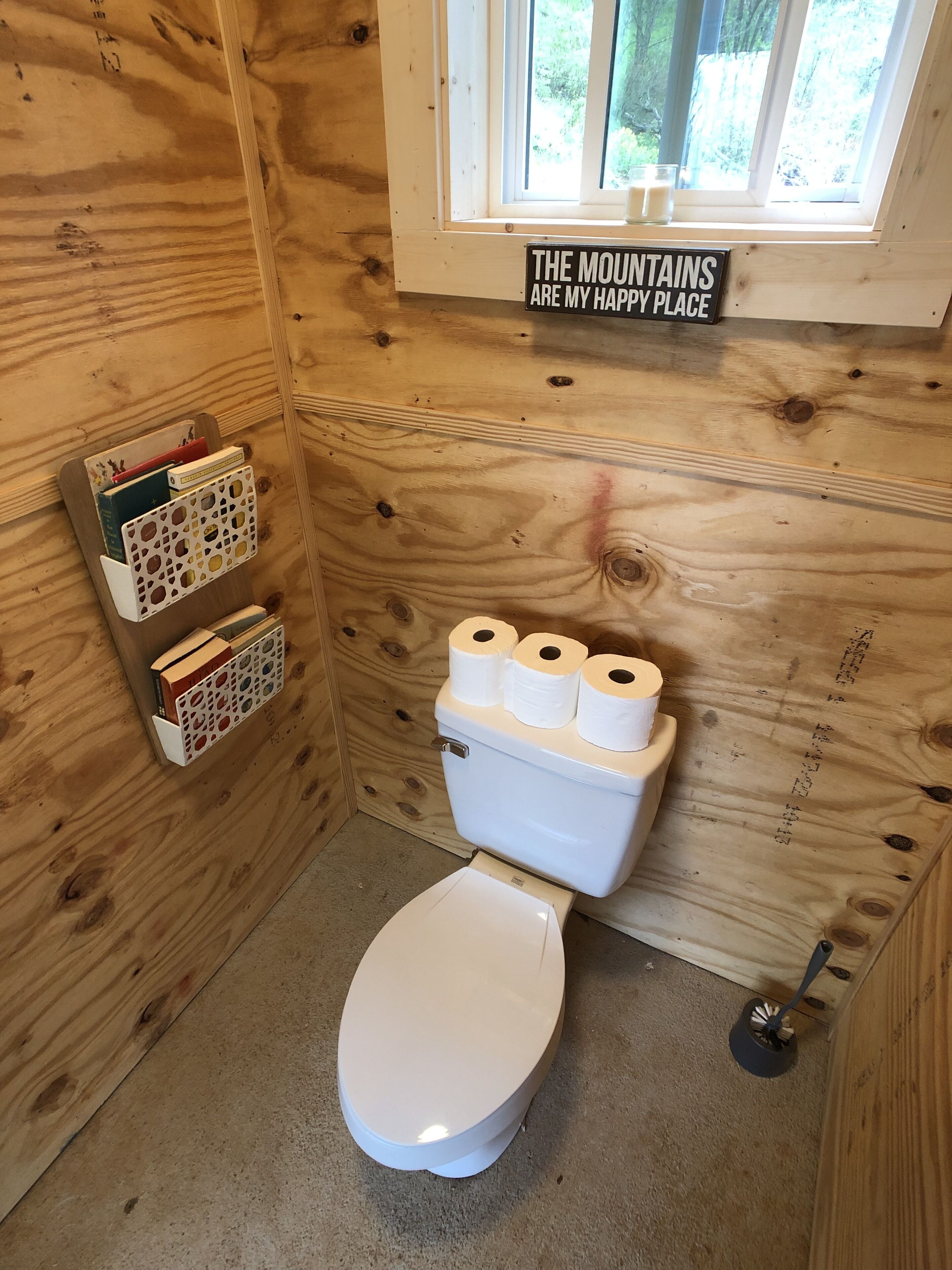 A wooden cabin bathroom with a toilet, a toilet paper holder, and a sign above the window that reads "THE MOUNTAINS ARE MY HAPPY PLACE".