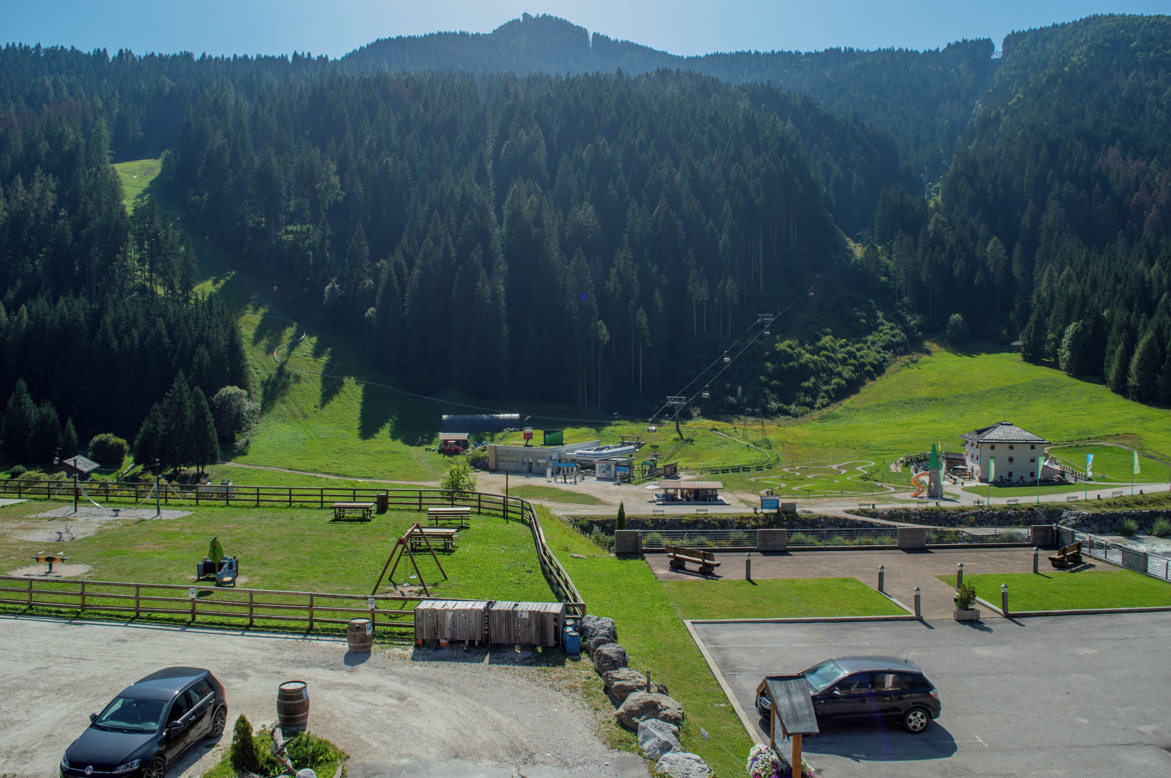 A rural setting with a parking area, a small building, and a fenced area with a playground.