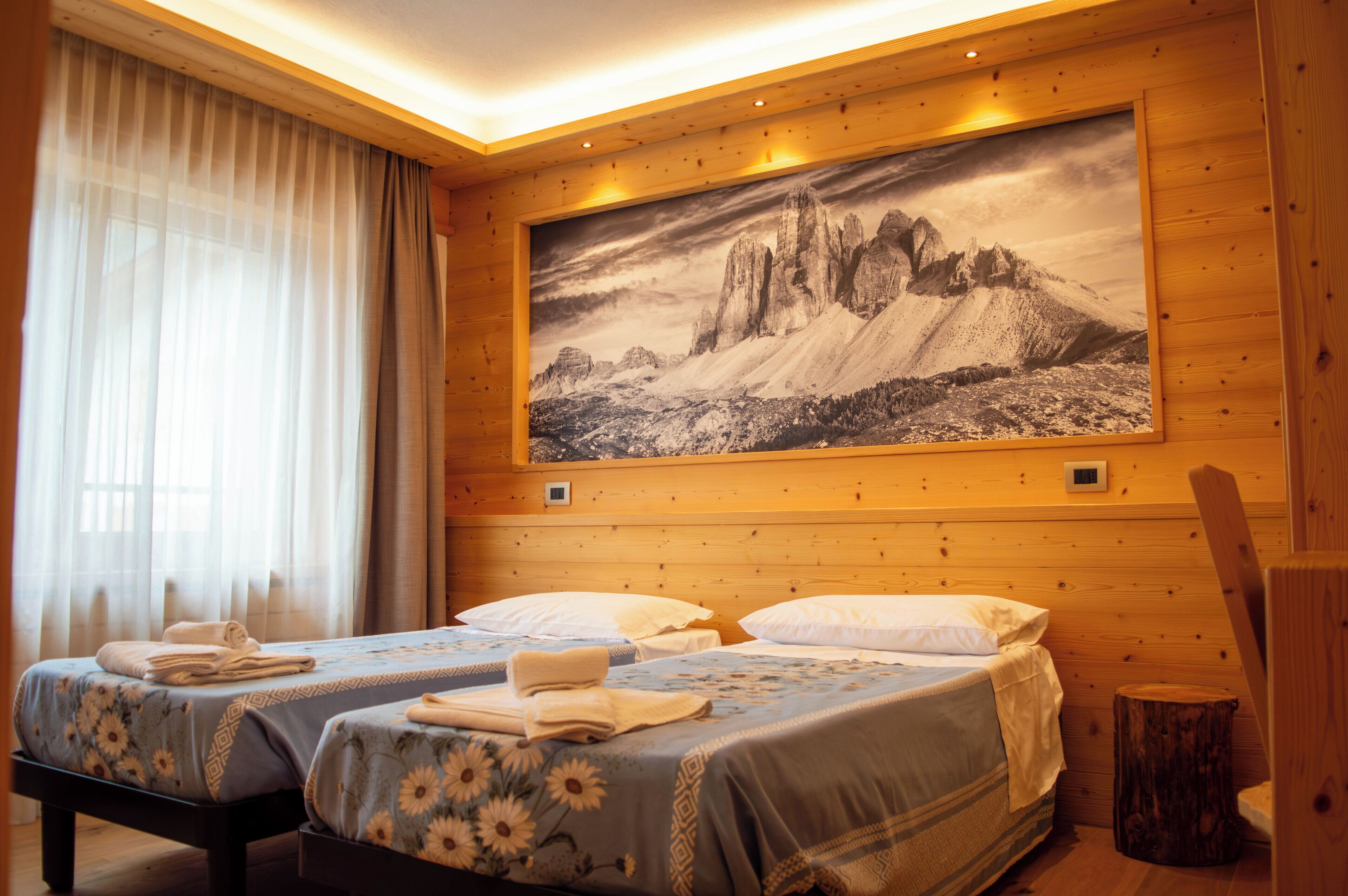 Une chambre d’hôtel avec deux lits, un grand tableau représentant un paysage de montagne accroché au mur, des lambris en bois et une fenêtre ornée de rideaux légers.