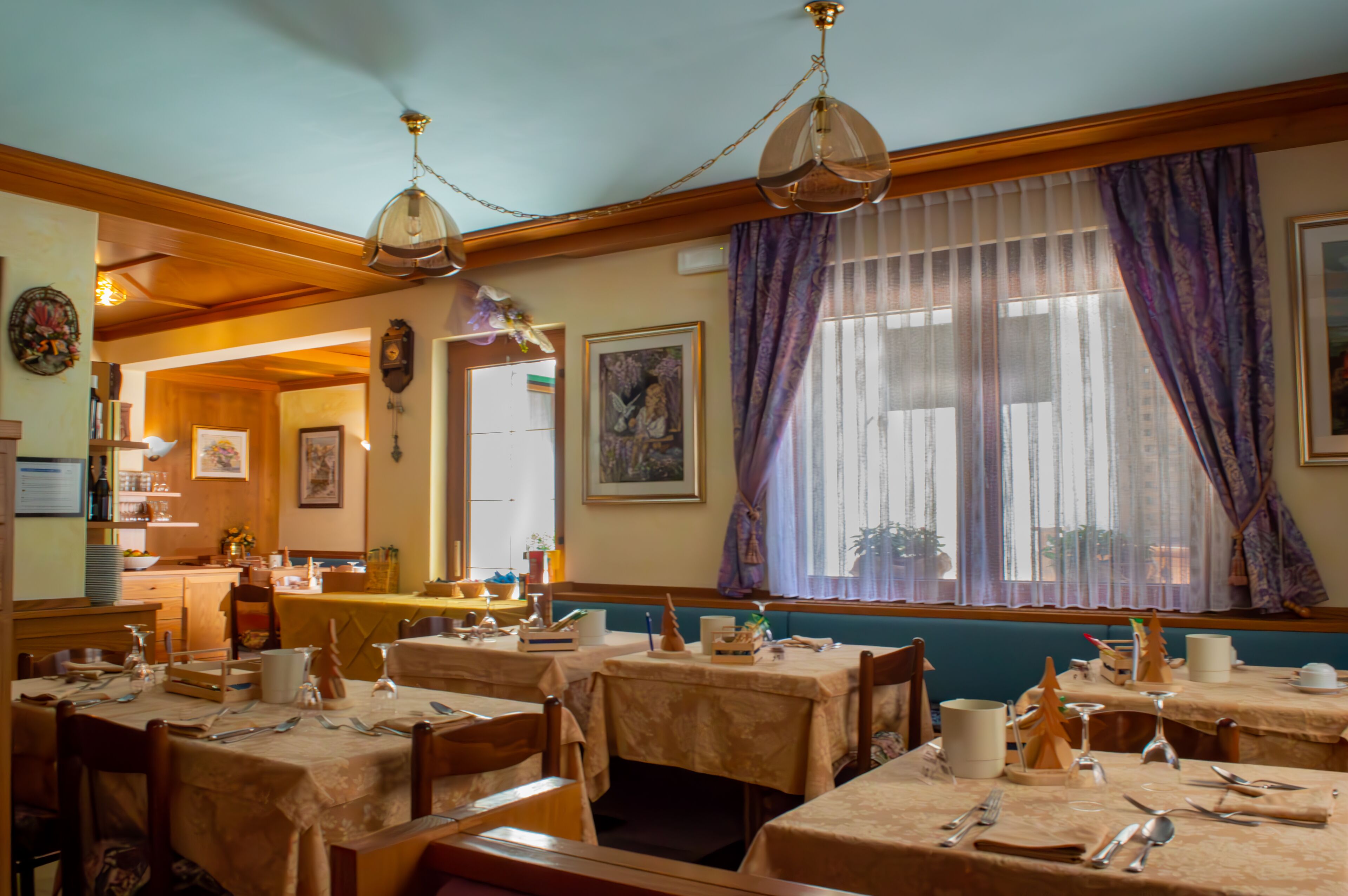 Restaurante com várias mesas preparadas para refeições, vigas de madeira no teto e uma grande janela com cortinas roxas.