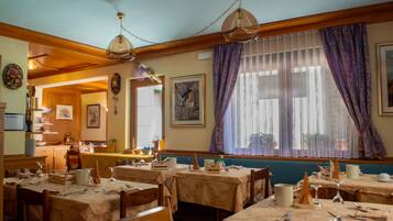 Restaurante com várias mesas preparadas para refeições, vigas de madeira no teto e uma grande janela com cortinas roxas.