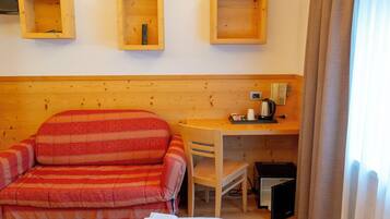A hotel room with a bed, a striped armchair, a desk with a telephone and a kettle, and wooden shelves above the bed.