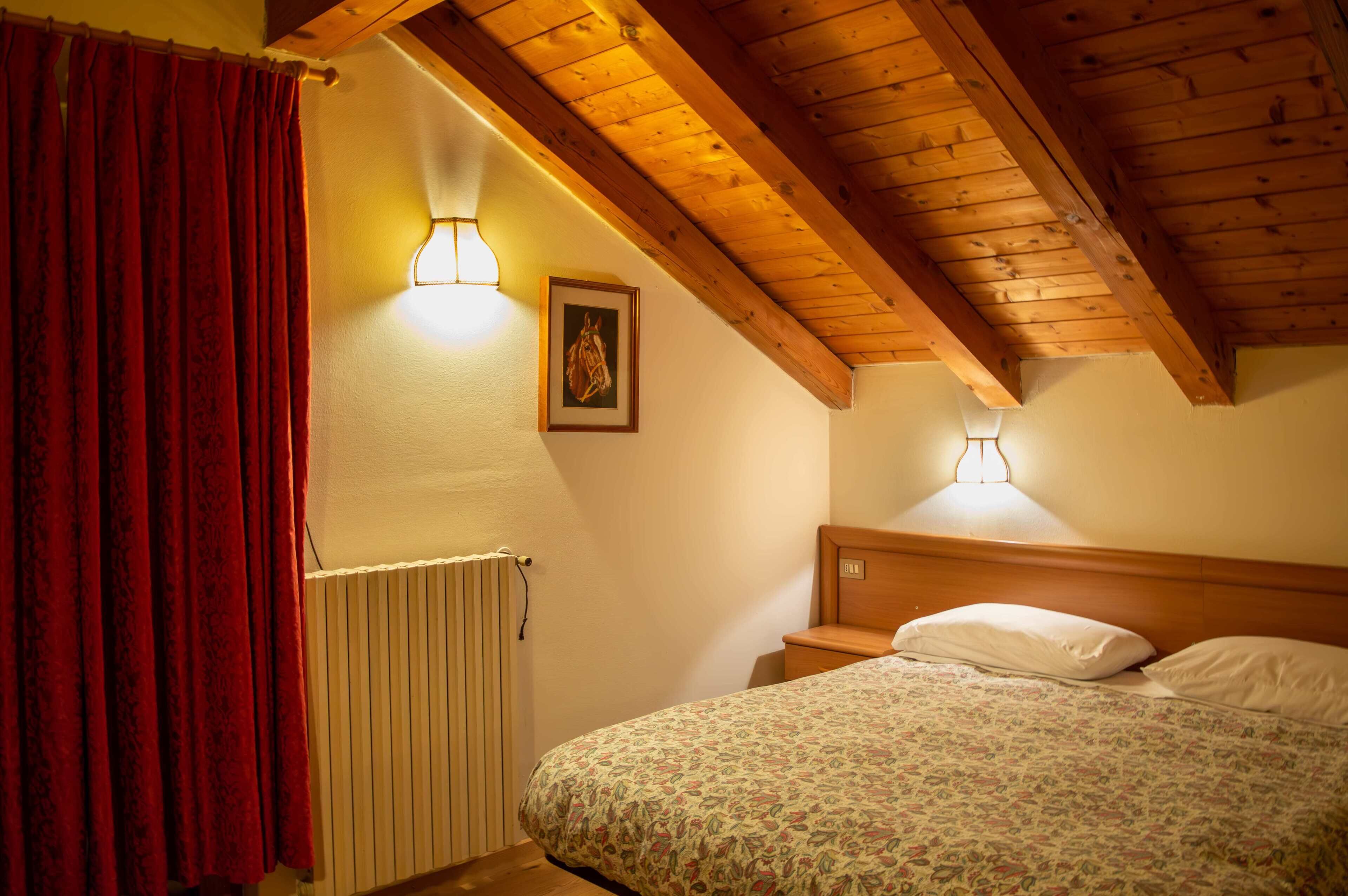 A bedroom with a wooden ceiling, a radiator, a bed with a floral bedspread, and two wall-mounted lamps.