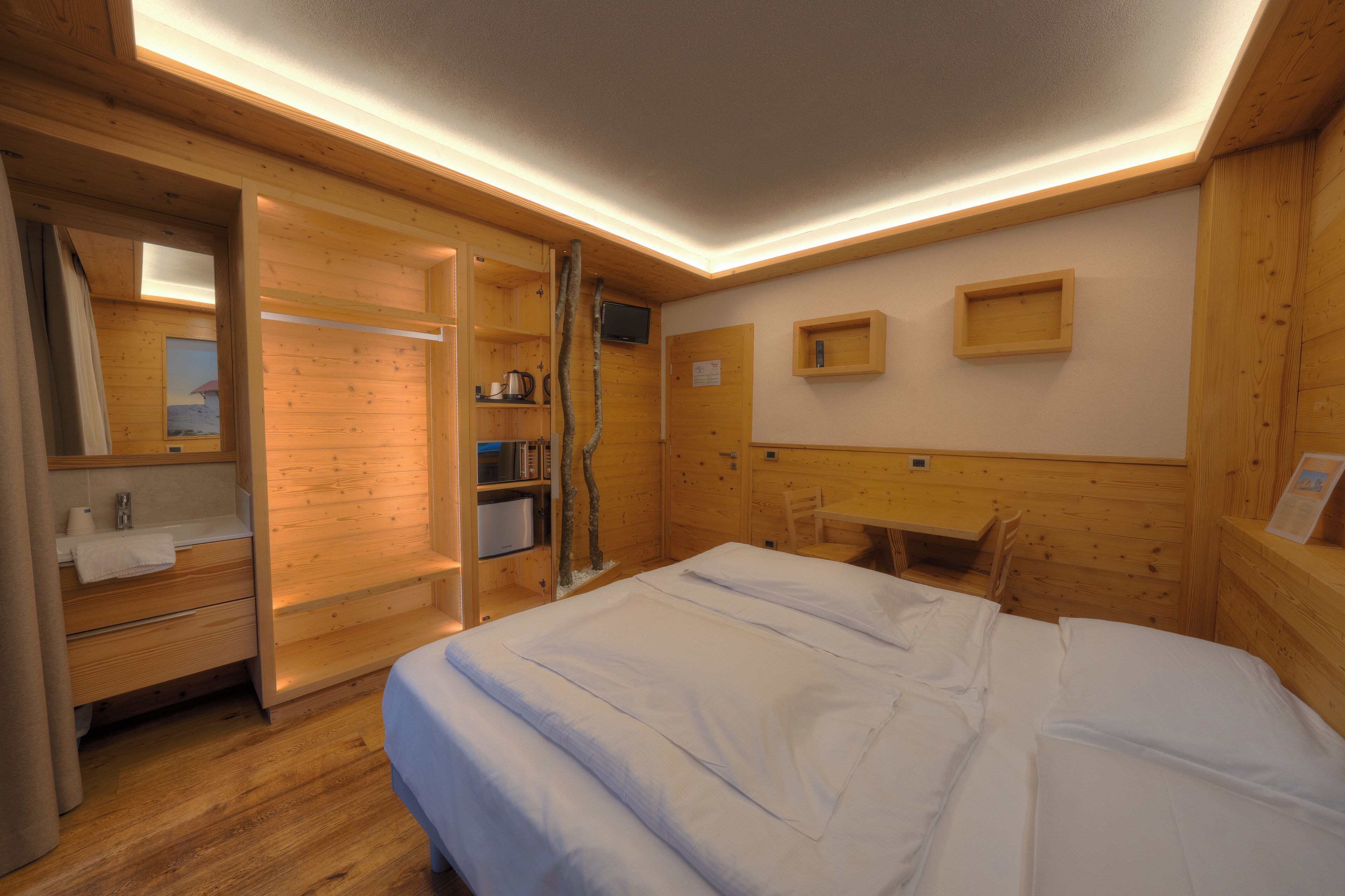 Una habitación de madera con una cama, un escritorio, una silla, un espejo y una repisa con una tetera.