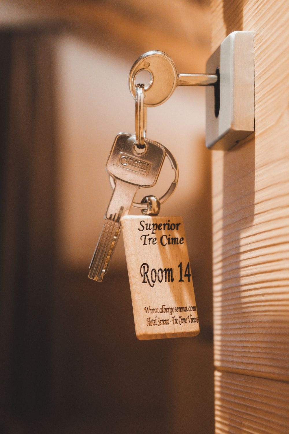 A keychain with a wooden tag attached to a key, hanging from a keyhole. The tag reads "Superior Tre Cime Room 14" and includes a website URL.