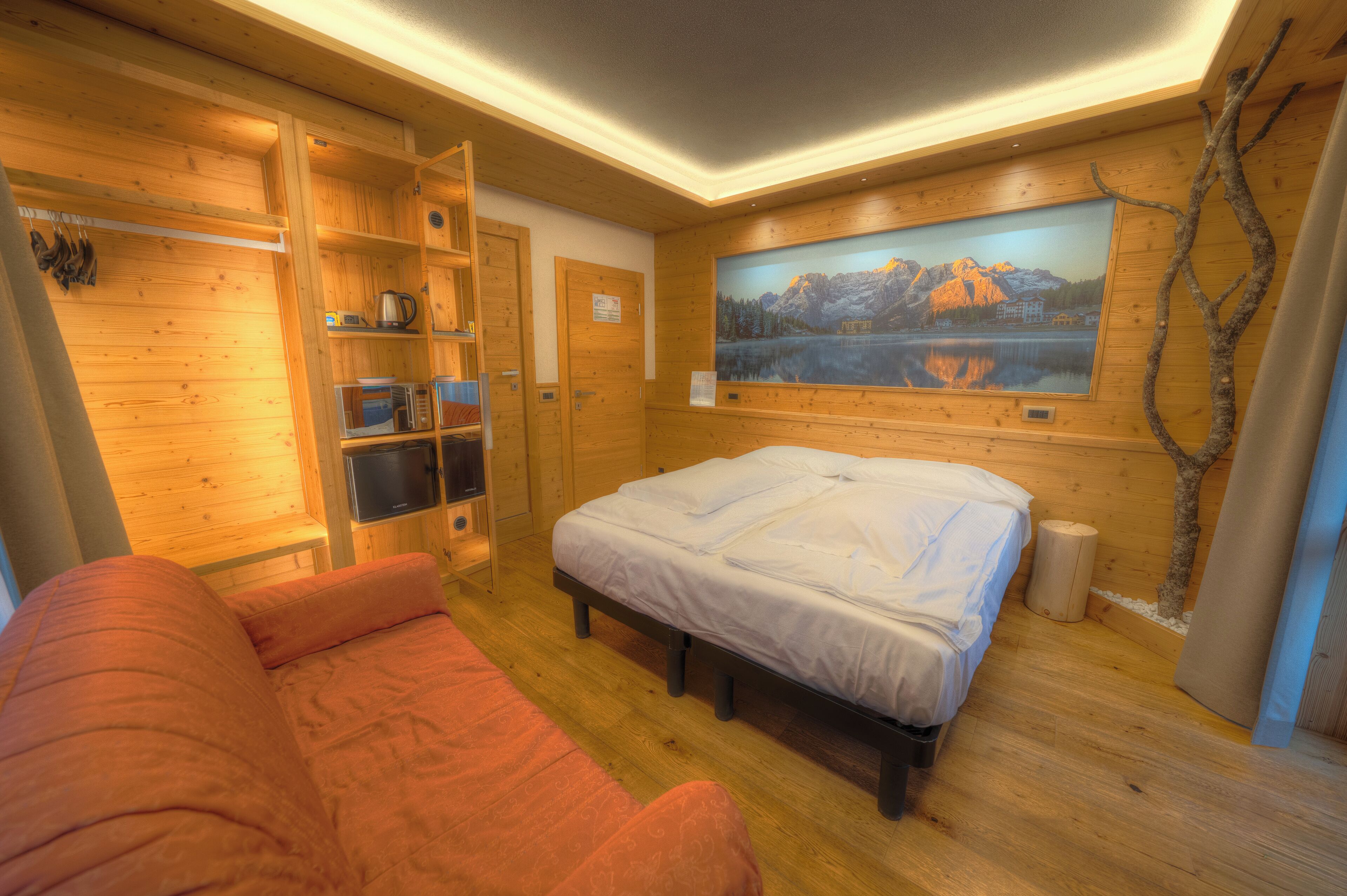 A wooden cabin room with a bed, a sofa, a painting of mountains, and a wooden shelf with kitchen items.