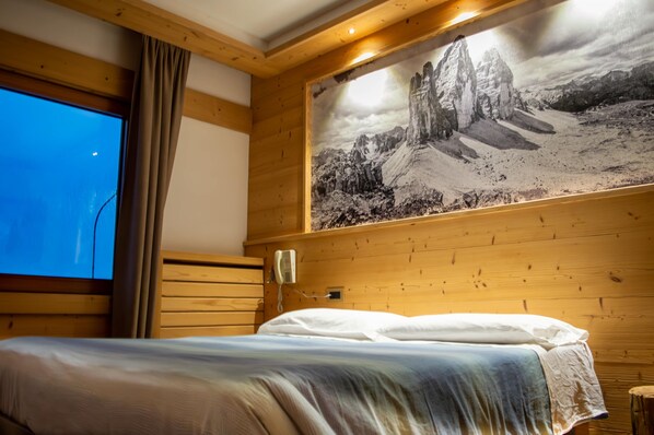 Una habitación de madera con una cama, un ventanal grande, un teléfono y un cuadro de un paisaje montañoso en la pared.