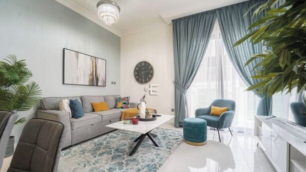 A modern living room with a grey sofa, a coffee table, and a blue armchair. There is a clock on the wall and a plant in the corner. - A modern luxurious 3 bedroom VILLA For family Gatherings, and Buiness meetings (AKOYA 2 - Damac Hills)