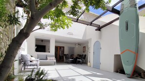A sunlit patio with a white sofa, dining table, and chairs, surrounded by greenery and a surfboard. - Charming seaside house with home office (Pag)