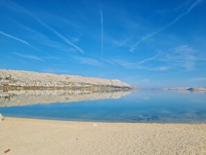 A calm lake with a clear sky, a sandy shore, and a distant mountain range. - Charming seaside house with home office (Pag)