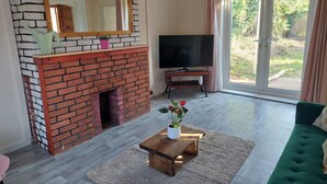 Uma sala de estar com lareira de tijolos, televisão sobre um suporte de madeira, sofá verde e vista para o exterior através de grandes janelas.