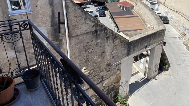Un balcon doté d’une balustrade en métal, de plantes en pot et offrant une vue sur une rue bordée de voitures garées et d’immeubles.
