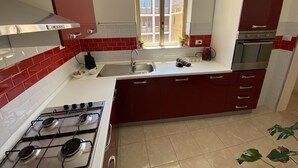 Une cuisine avec des carreaux rouges et blancs, une cuisinière à gaz, un évier et une fenêtre donnant sur les bâtiments environnants.