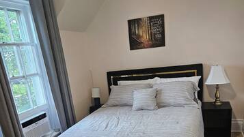A bedroom with a bed, a nightstand, a lamp, a window with curtains, a radiator, and a painting on the wall.
