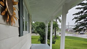 A covered porch with a wooden floor, white columns, and a sunflower decoration on the wall. - Sunflower Guest Suites (Summerside)