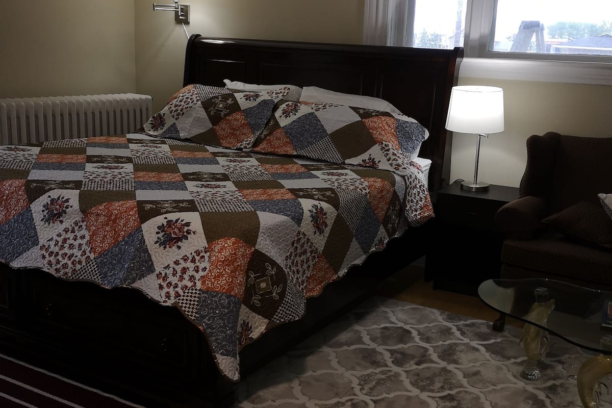 A bedroom with a bed, a nightstand, a lamp, a chair, and a window with blinds.