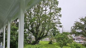 A covered porch with white columns, a grassy lawn, and a large tree. - Sunflower Guest Suites (Summerside)