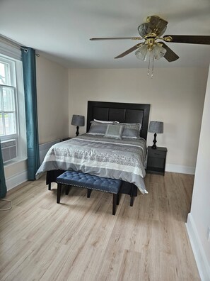 A bedroom with a bed, two bedside tables, a bench, a ceiling fan, and a window with curtains. - Sunflower Guest Suites (Summerside)