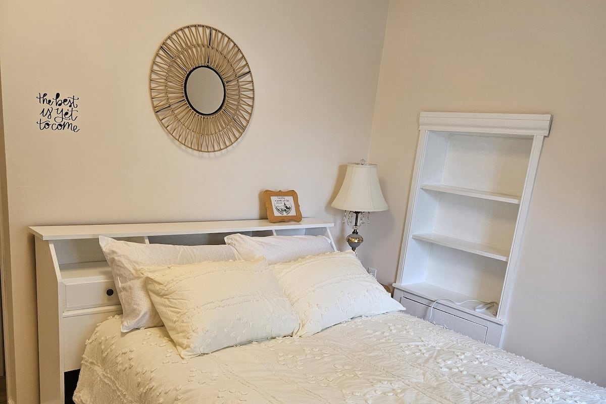 A neatly made bed with a white duvet and pillows, a bedside table with a lamp, a wall mirror, and a shelf with a framed picture.