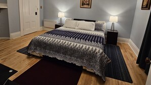 A bedroom with a bed, two bedside tables, a chandelier, a door, and a framed picture on the wall. - Sunflower Guest Suites (Summerside)