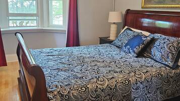 A bedroom with a large bed, a nightstand, a lamp, a window with curtains, and a painting on the wall.